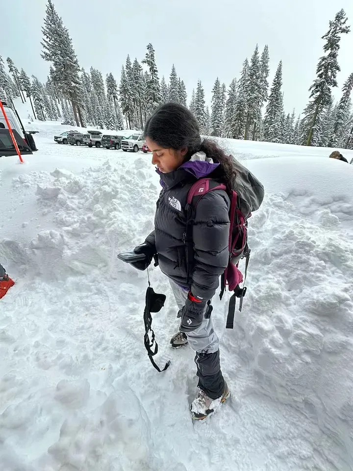 Ananya in a down jacket and crampons walking through deep snow beside a parking lot