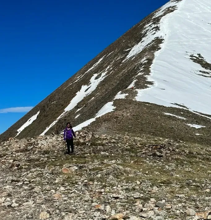 Ananya in a purple jacket standing at the foot of a steep, snow-streaked ridge