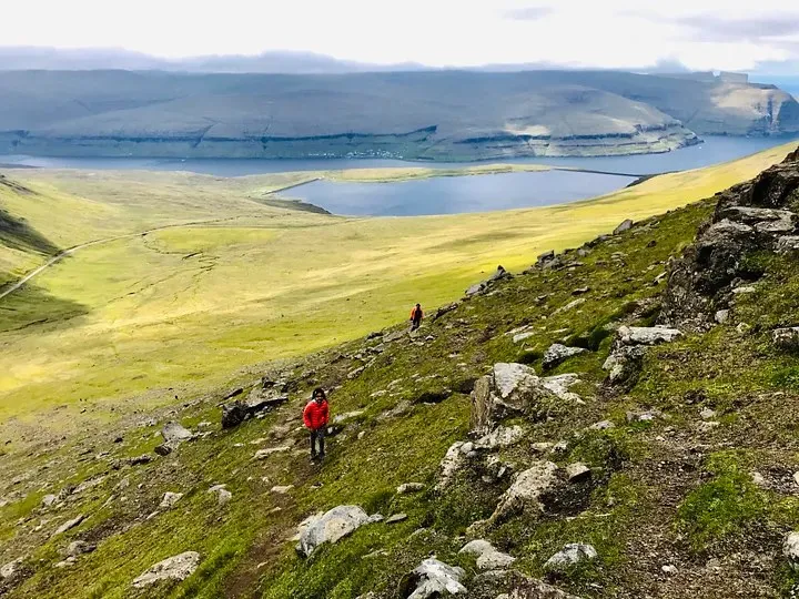 Ananya in a red jacket on a steep, rocky grass slope above a fjord and lake
