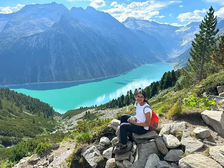 Ananya sitting on rocks above the turquoise Schlegeis Reservoir, with mountains beyond