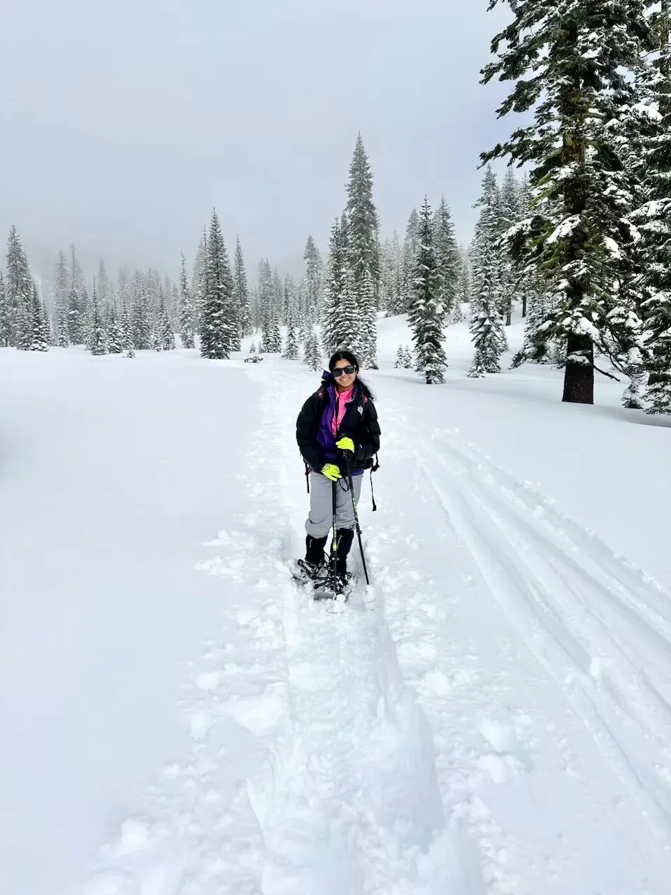 Ananya smiling on snowshoes with trekking poles on a snowy trail among pine trees